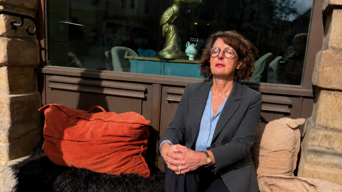„Eine Frau mittleren Alters mit lockigem braunem Haar und Brille sitzt entspannt mit geschlossenen Augen und gefalteten Händen vor einem Café und entspannt.